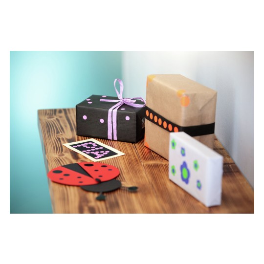 De afbeelding toont een houten tafel met ingepakte cadeaus in verschillende kleuren en een kaart met een stippenpatroon. Er is ook een papieren lieveheersbeestje gemaakt van kleurpapier.