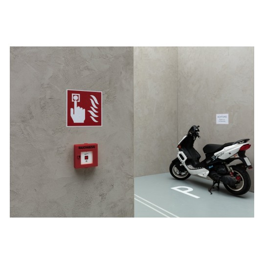 De afbeelding toont een muur met een brandveiligheidsbord en een rookafzuiging. Daarnaast staat een motorfiets geparkeerd in een speciale ruimte.