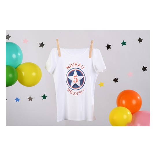Een witte t-shirt met de tekst 'Niveau 5 Reussi!' en sterren eromheen, naast kleurrijke ballonnen.