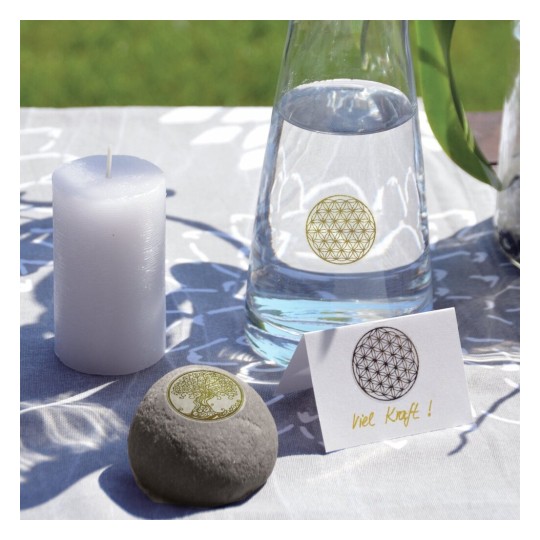 Op de afbeelding staat een tafel met een witte kaars, een karaf water, en een grijze steen met een boomontwerp. Daarnaast ligt een kaartje met de tekst 'Veel Kracht!' en een cirkelontwerp. De tafelkleed is lichtgrijs met patronen.