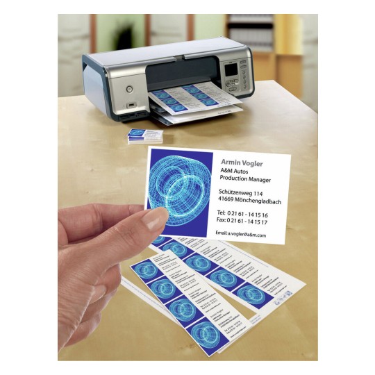In de afbeelding is een printer zichtbaar die visitekaartjes afdrukt. Een hand houdt een visitekaartje vast met contactinformatie en een grafisch ontwerp. Het lijkt een kantoorsetting.