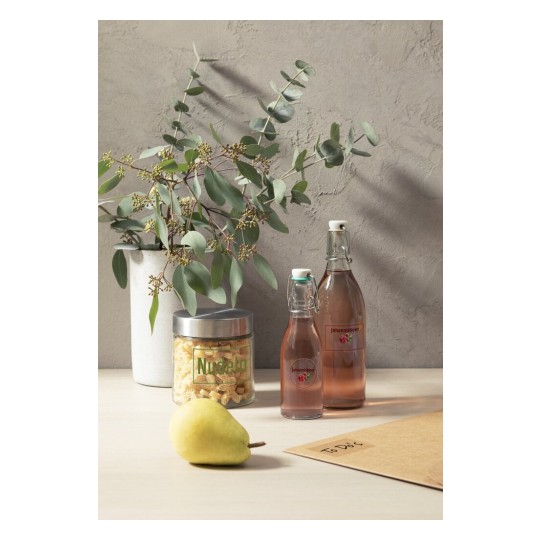 De afbeelding toont een moderne tafelsetting met een vaas eucalyptus, twee flessen met johannisbeeren, een doos met noedels en een peer.