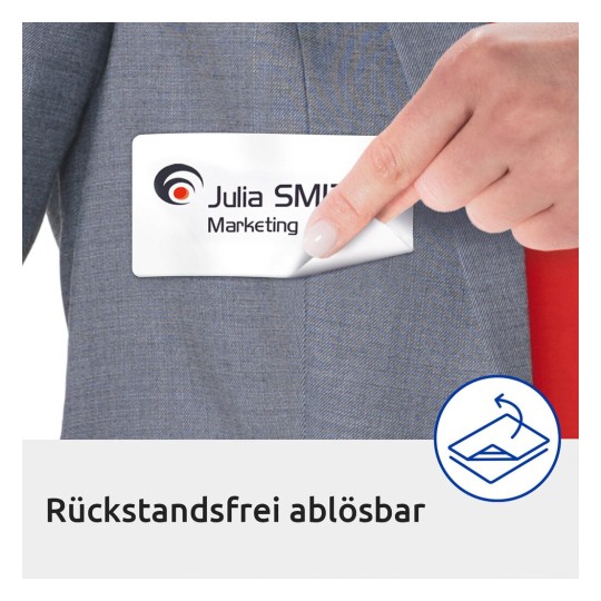 In de afbeelding ziet u een hand die een label uit een zak van een grijze jas trekt. Het label heeft de naam 'Julia SMITH Marketing' en de tekst 'Rückstandsfrei ablösbar', wat betekent dat het vrij kan worden verwijderd zonder resten achter te laten.