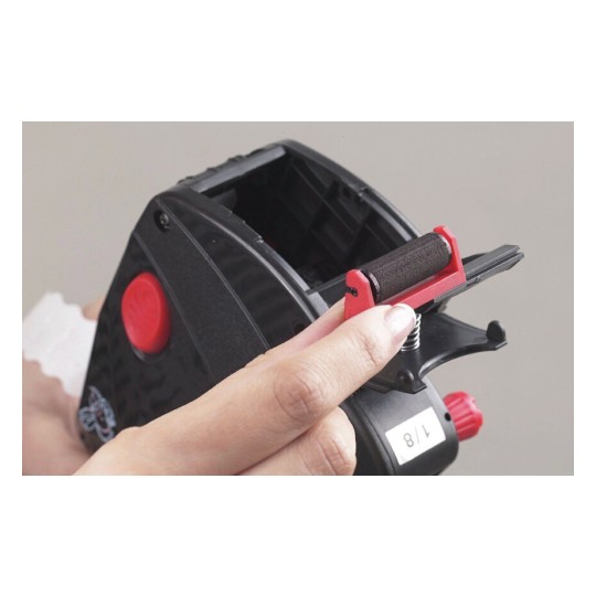 Dit is een hand die een onderdeel vasthoudt van een apparaat dat lijkt op een etiketteermachine. Het onderdeel is rood en zwart, met een veermechanisme, en het apparaat is voornamelijk zwart.