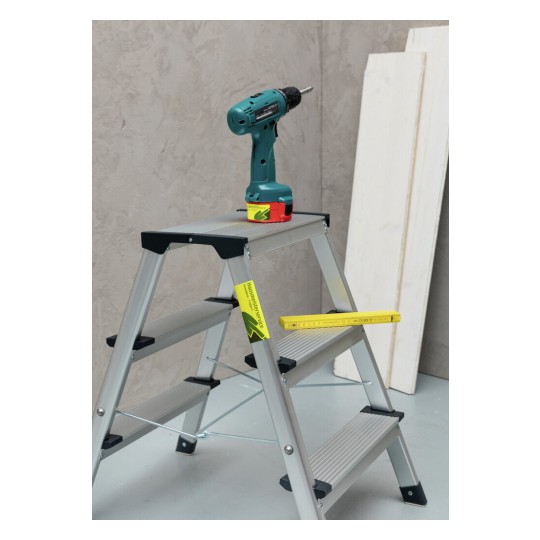 De afbeelding toont een ladder met een boormachine erop, naast enkele houten planken. De ladder is van aluminium en heeft een meetlat erop liggen.