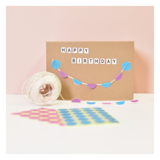 De afbeelding toont een verjaardagskaart met de tekst 'HAPPY BIRTHDAY' erop en feestelijke decoraties in de vorm van gekleurde cirkels. Daarnaast ligt een bolletje touw en kleurrijke stickervellen op de tafel.