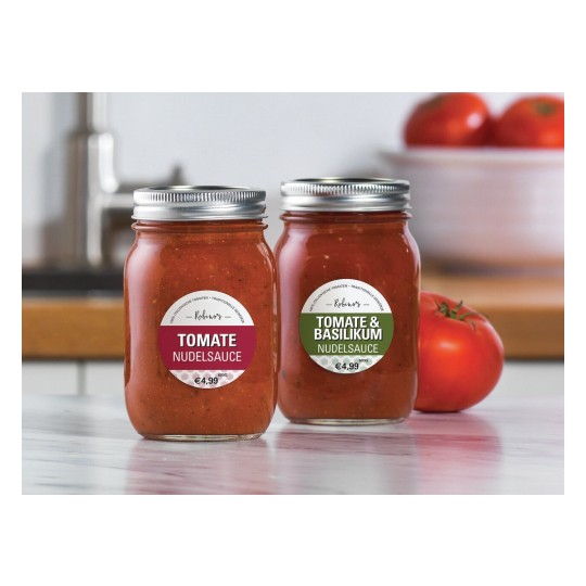De afbeelding toont twee glazen potten met pastasaus. De linker pot heeft het etiket 'Tomate Noodlesaus' en de rechter pot heeft het etiket 'Tomate & Basilikum Noodlesaus'. Op de achtergrond zijn enkele tomaten te zien.