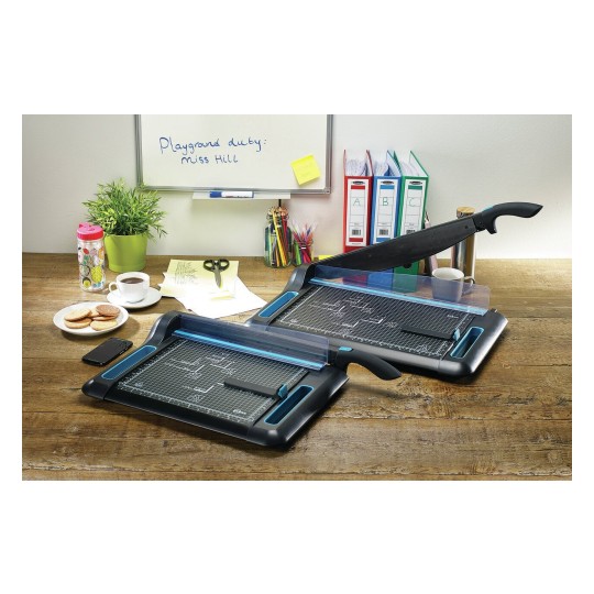 Het beeld toont een bureau met twee snijmachines, een plantenpot, een beker koffie, koekjes, een mobiele telefoon, scharen, gekleurde pennen en mappen. Er is ook een whiteboard met een boodschap.