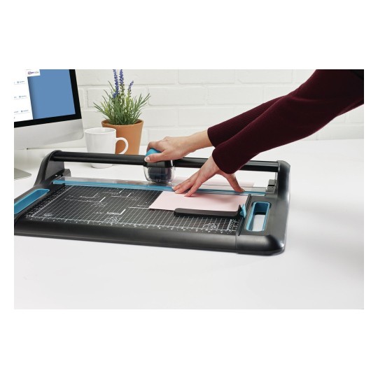 Een persoon gebruikt een snijmachine op een bureau. Op de achtergrond staat een computer, een witte mok en een pot met planten.