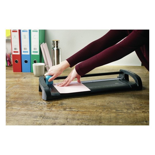 Een hand snijdt papier met een snijmachine op een houten tafel. Op de achtergrond staan ordners en een mok.