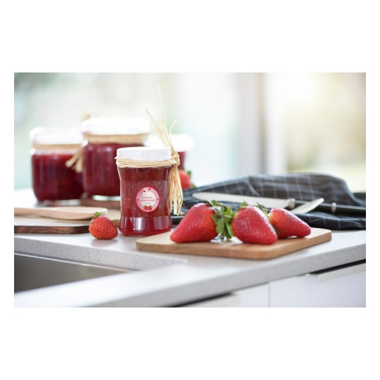 De foto toont een keuken met aardbeienjam in glazen potten, verse aardbeien en houten snijplanken.