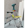 De afbeelding toont een ladder met een boormachine erop, naast enkele houten planken. De ladder is van aluminium en heeft een meetlat erop liggen.