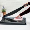 Een hand snijdt een roze vel papier met een snijmal terwijl een plant en een kopje op de achtergrond zichtbaar zijn.