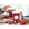 Op de afbeelding zijn glazen potten met aardbeienjam te zien. Een hand plaatst een label op een pot, terwijl er verse aardbeien op de tafel liggen. De potten zijn versierd met een strakdeksel en een natuurlijke touwbinding voor een huiselijke uitstraling.