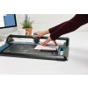 Een persoon gebruikt een snijmachine op een bureau. Op de achtergrond staat een computer, een witte mok en een pot met planten.