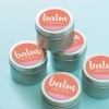De afbeelding toont een aantal metalen blikjes lippenbalsem, die gestapeld zijn. Elk blikje heeft een etiket met de tekst 'balm' en de vermelding '100% Natural Beeswax Lip Balm'. De blikjes zijn op een lichtblauwe achtergrond geplaatst.